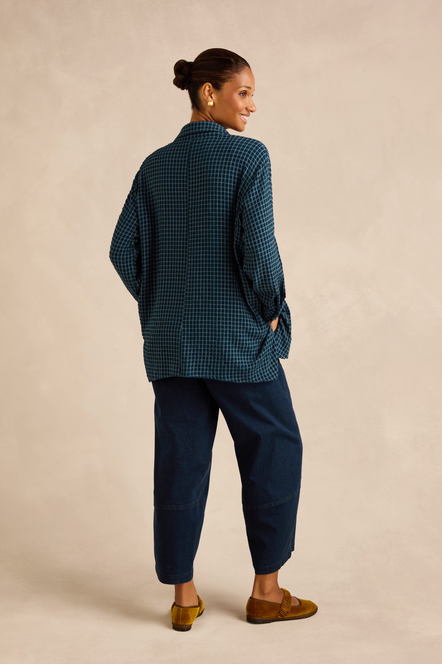 A woman stands facing away, wearing the Seersucker Squares Relaxed Shirt with a blue checkered pattern and boxy silhouette, paired with navy pants and brown shoes, her hair in a bun against a neutral background.