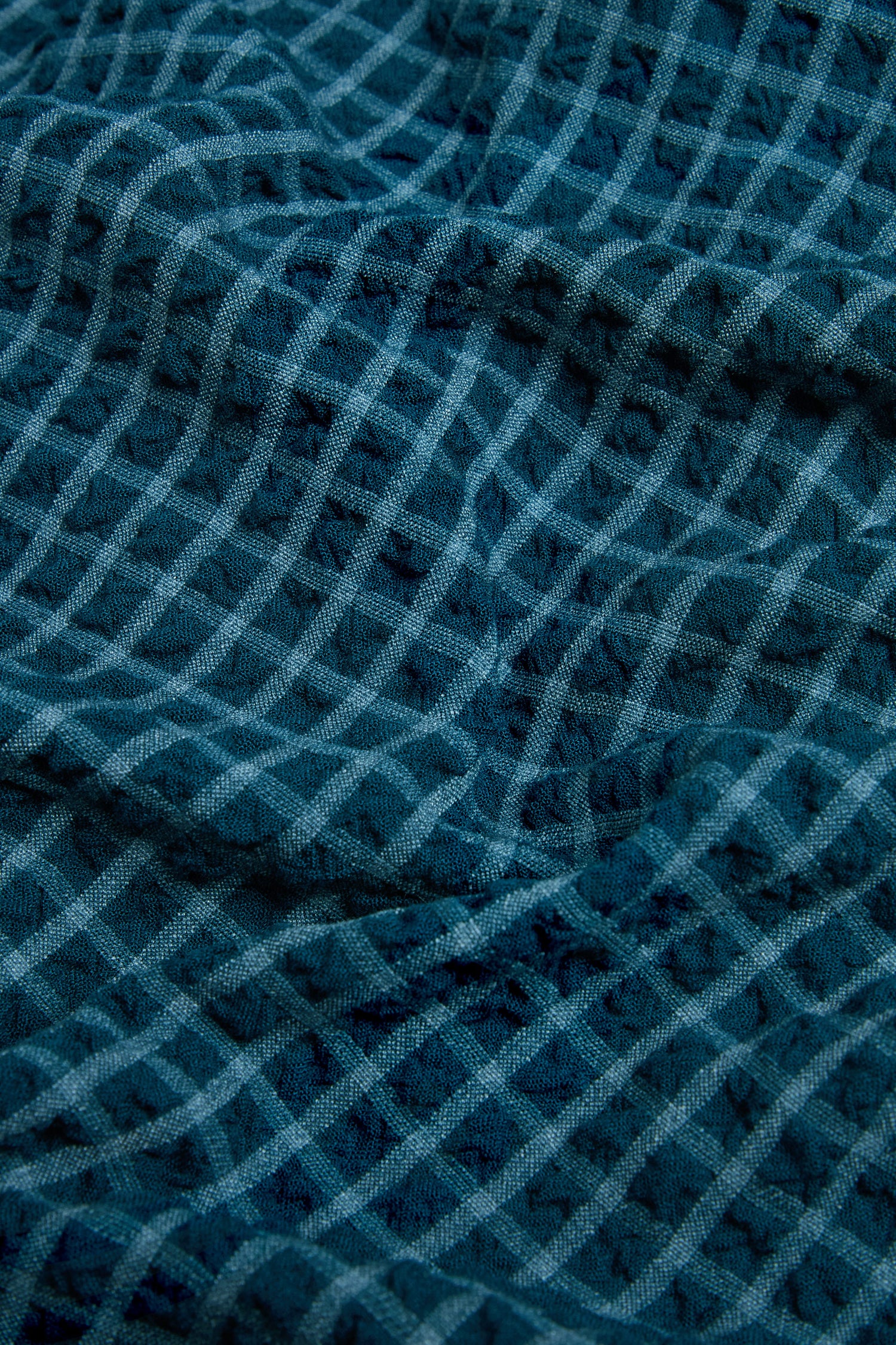 Close-up of blue fabric with a repeating grid pattern, highlighting the texture and subtle folds that characterize the Seersucker Squares Relaxed Shirt—crafted in England with a breathable weave and signature boxy silhouette.