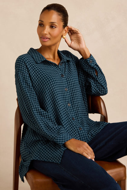 A woman sits on a chair wearing the Seersucker Squares Relaxed Shirt, which features a blue checkered pattern and a boxy silhouette, paired with dark pants. She touches her ear with one hand and gazes to the side.