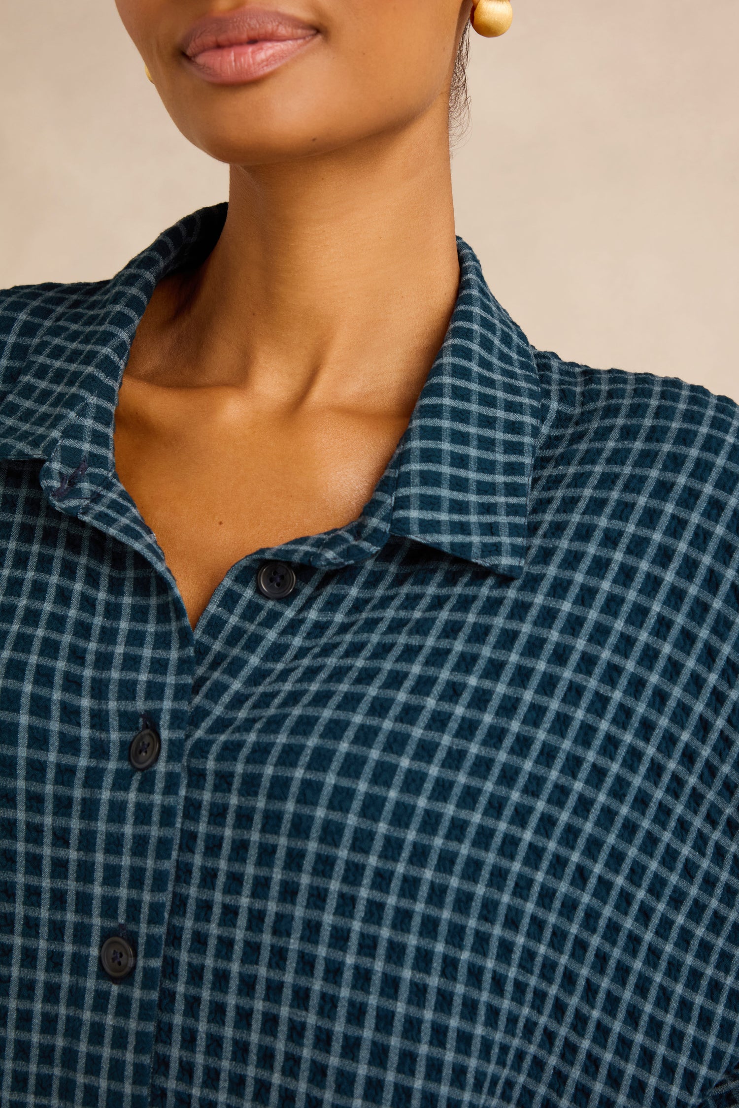 A person wearing the Seersucker Squares Relaxed Shirt in a blue and black checkered pattern, styled with gold round earrings, pictured from the shoulders up.
