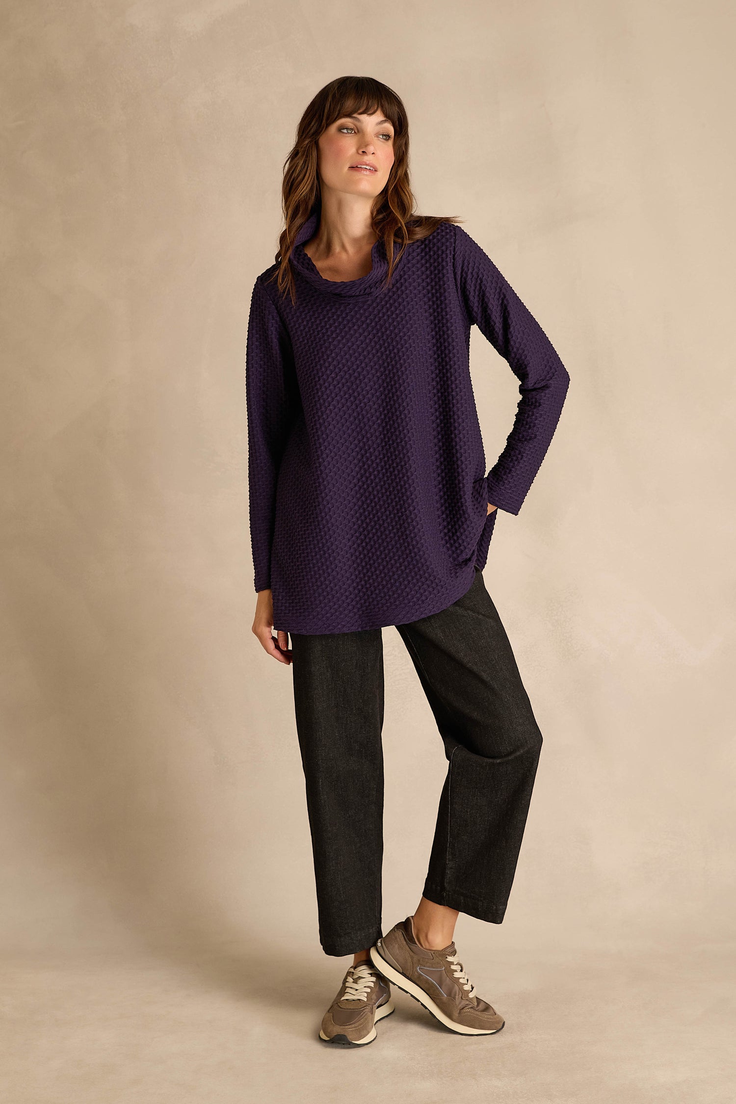 A woman stands against a beige backdrop wearing the Waffle Textured Jersey Damson Cowl Neck Top, paired with dark wide-leg pants and taupe sneakers, one hand in her pocket and a relaxed posture.