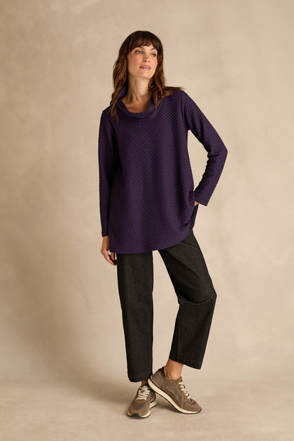 A woman stands against a beige backdrop wearing the Waffle Textured Jersey Damson Cowl Neck Top, paired with dark wide-leg pants and taupe sneakers, one hand in her pocket and a relaxed posture.