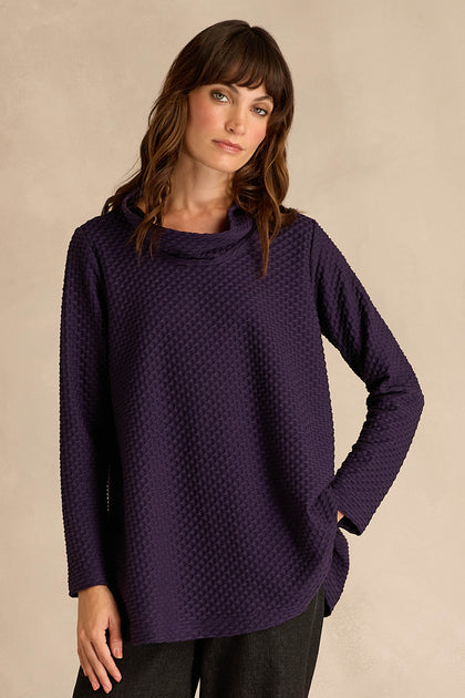 A woman stands against a plain beige background with one hand in her pocket, wearing the Waffle Textured Jersey Damson Cowl Neck Top and dark pants.