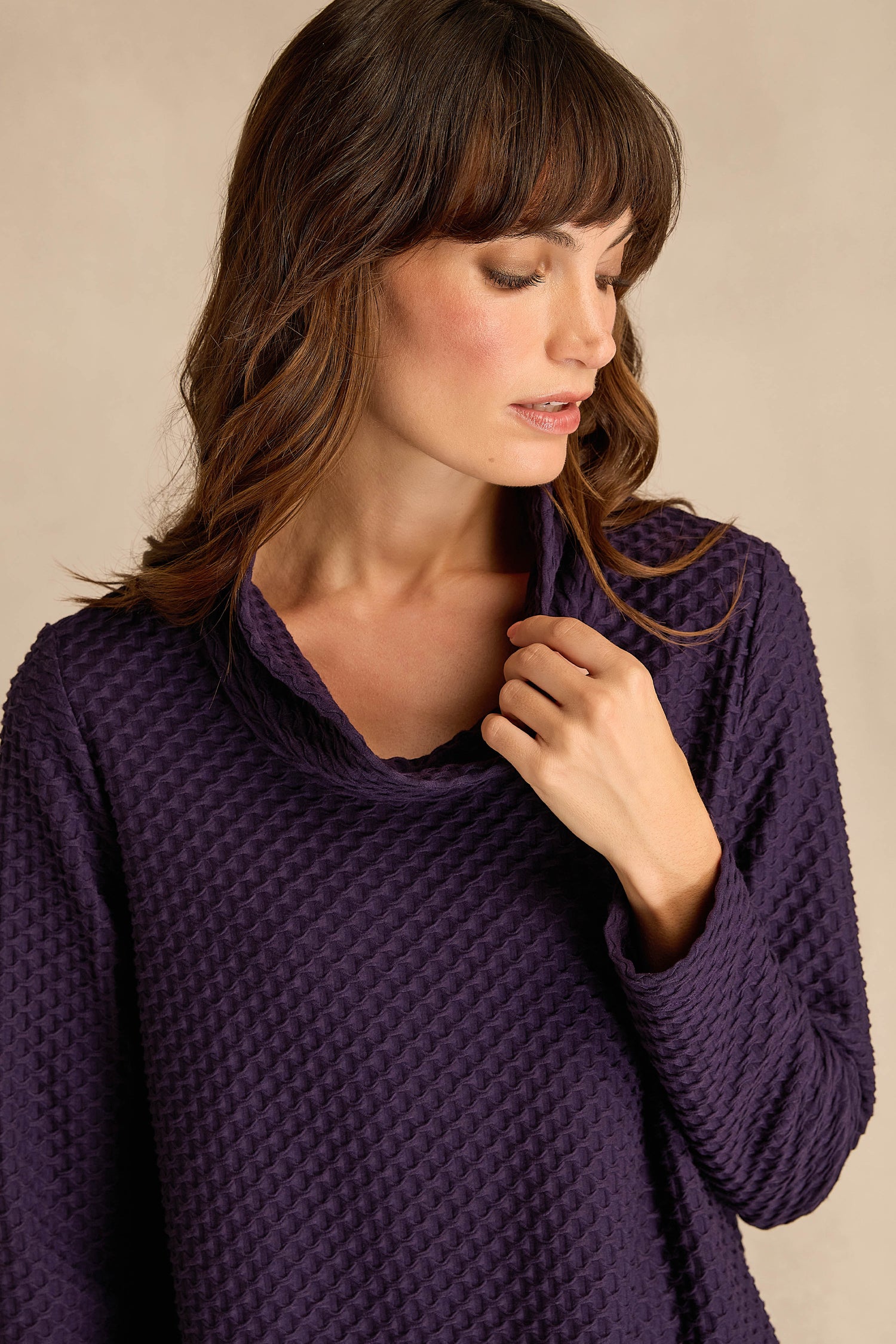 A woman wearing the Waffle Textured Jersey Damson Cowl Neck Top, a fit and flare design with long sleeves and a cowl neck, gently touches the collar as she looks down.