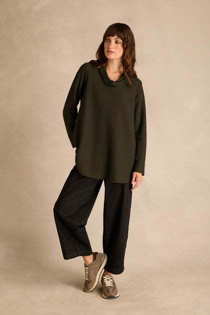 A woman stands against a neutral background, wearing the Waffle Textured Jersey Pine Cowl Neck Top, paired with wide-leg black pants and beige sneakers, with one hand in her pocket.