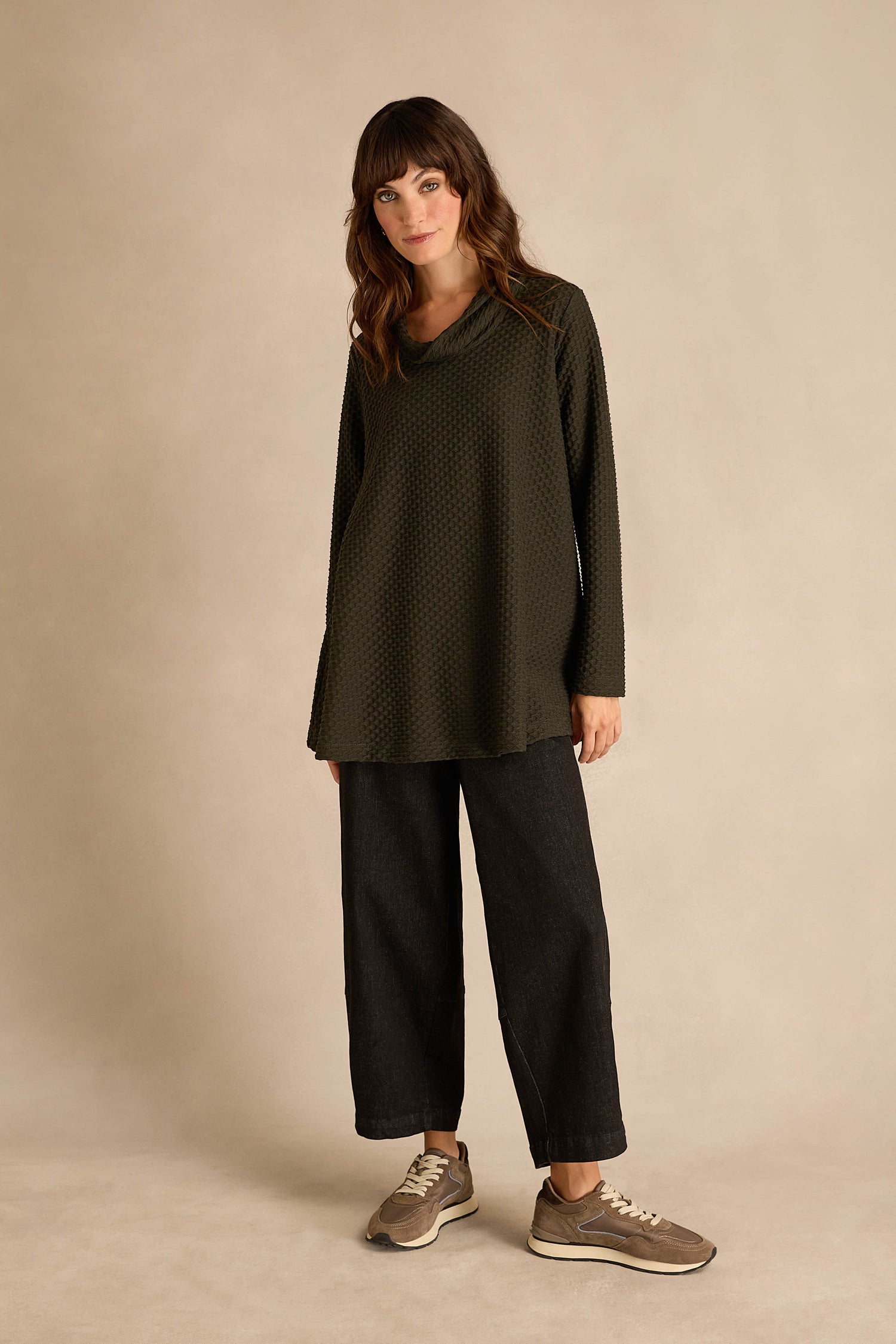 A woman stands in front of a beige background wearing the Waffle Textured Jersey Pine Cowl Neck Top, featuring loose long sleeves and a cowl neck, paired with wide-leg black pants and taupe sneakers. This top is made in England.