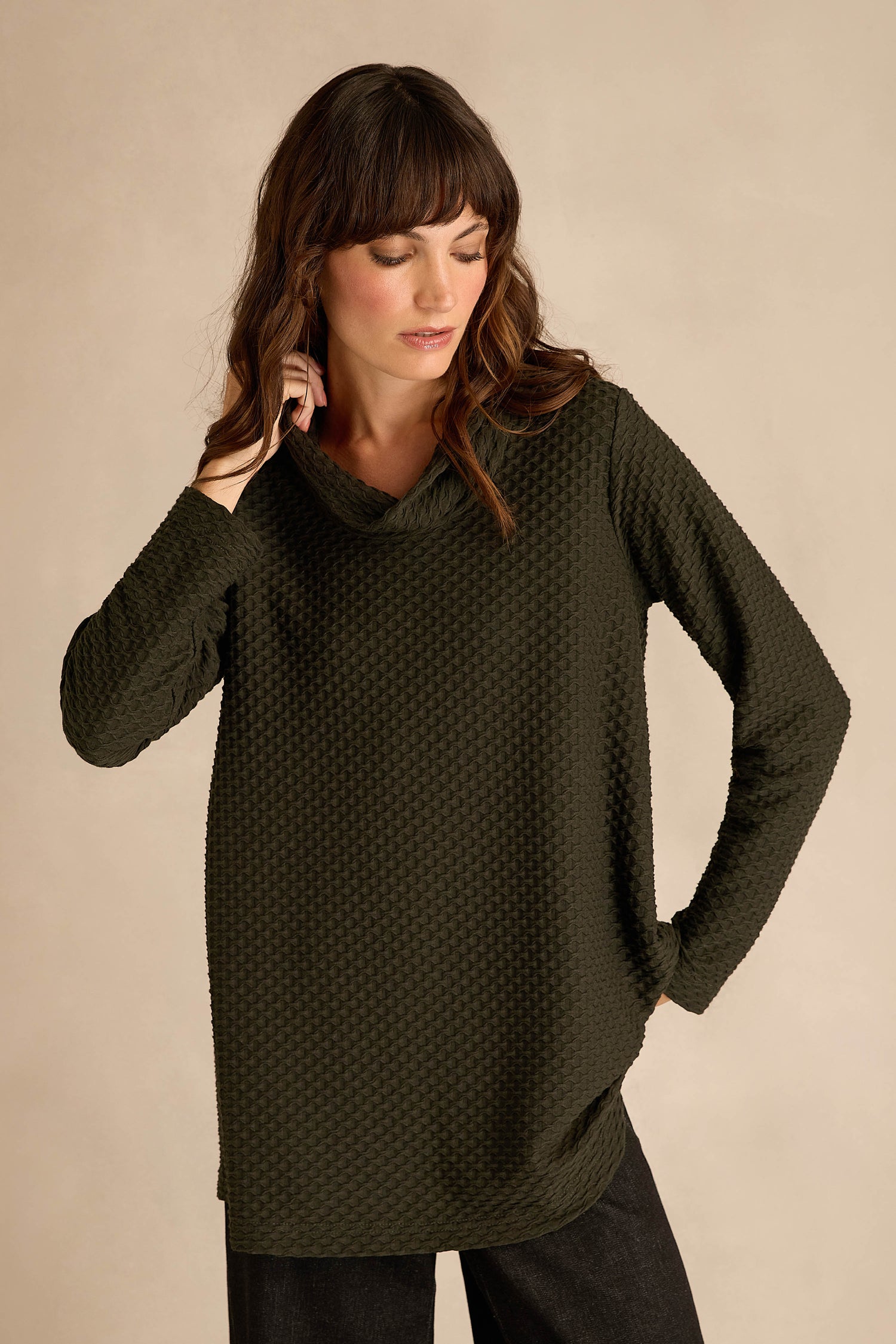 A woman stands against a plain beige background, looking downward, wearing the Waffle Textured Jersey Pine Cowl Neck Top—a long-sleeve, relaxed fit top in dark green with a distinctive waffle texture.
