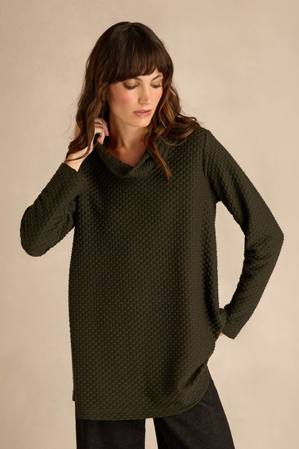 A woman stands against a plain beige background, looking downward, wearing the Waffle Textured Jersey Pine Cowl Neck Top—a long-sleeve, relaxed fit top in dark green with a distinctive waffle texture.