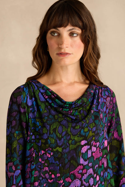 A woman with long brown hair wears the Winter Garden Jersey Cowl Neck Top, featuring a multicolored animal print and a draped neckline, as she stands against a neutral background.