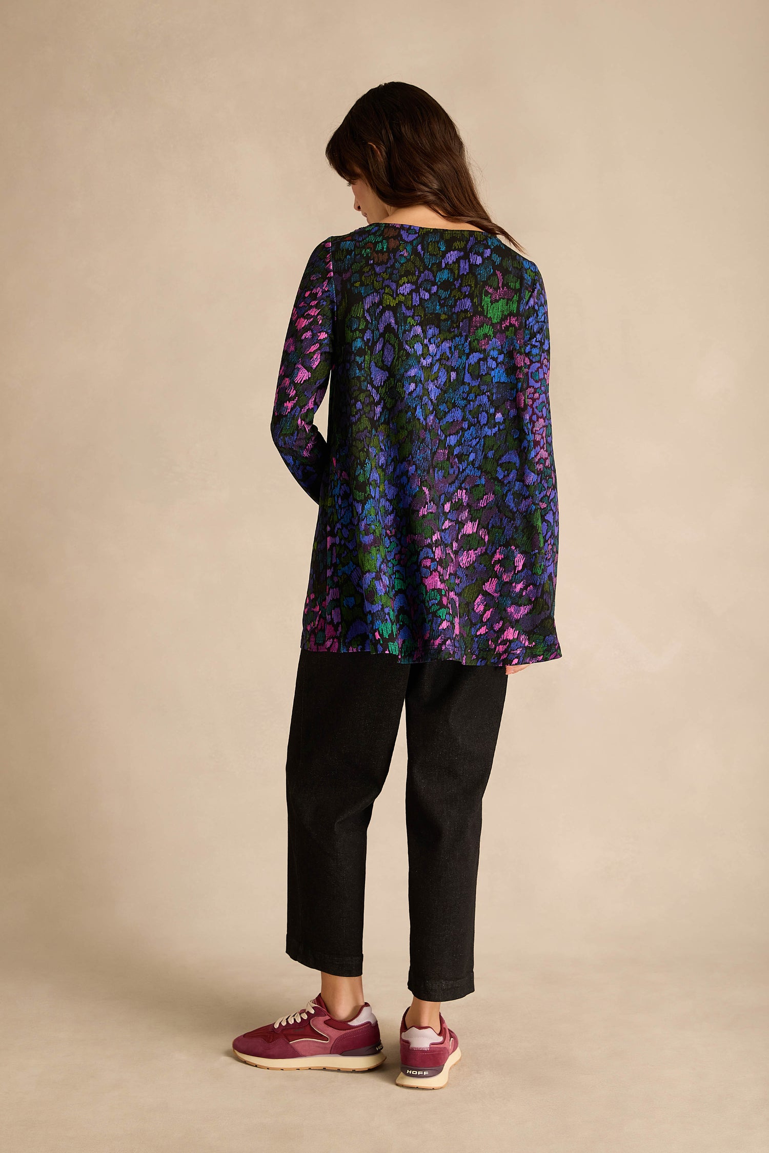 A person stands facing away against a neutral background, wearing the Winter Garden Jersey Cowl Neck Top—a long-sleeve, multicolored floral fit and flare top—paired with black pants and maroon sneakers.