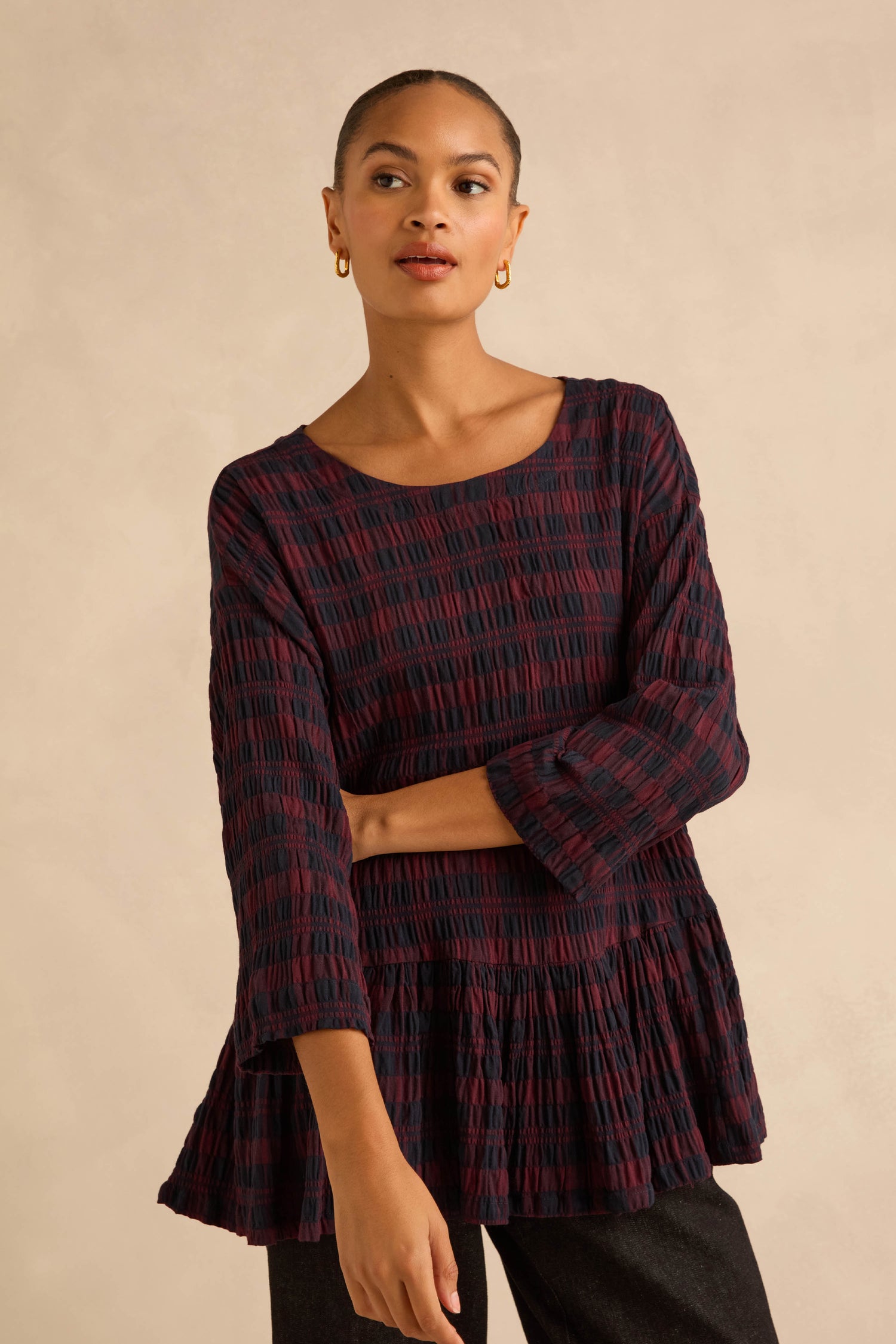 A person wearing a dark red and black Crinkle Check Frill Hem Top with long sleeves and a gathered peplum hem.