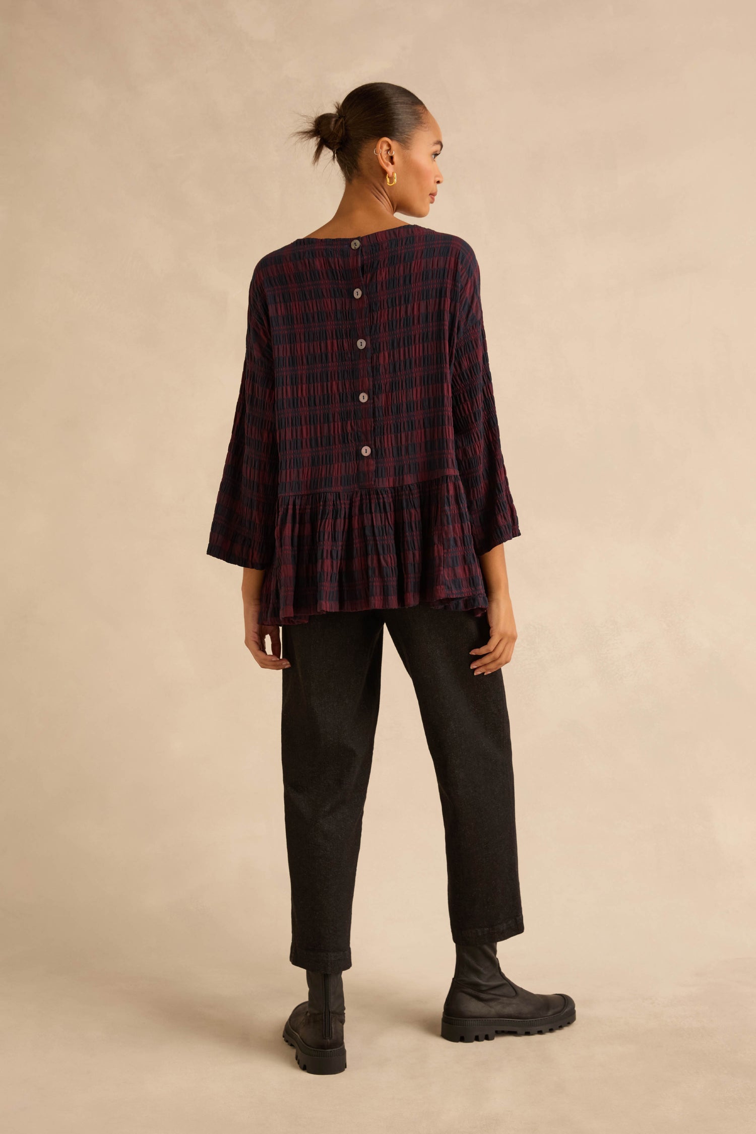 Back view of a person wearing a dark blue and red Crinkle Check Frill Hem Top with a peplum hem and buttoned back, paired with black trousers and black boots.