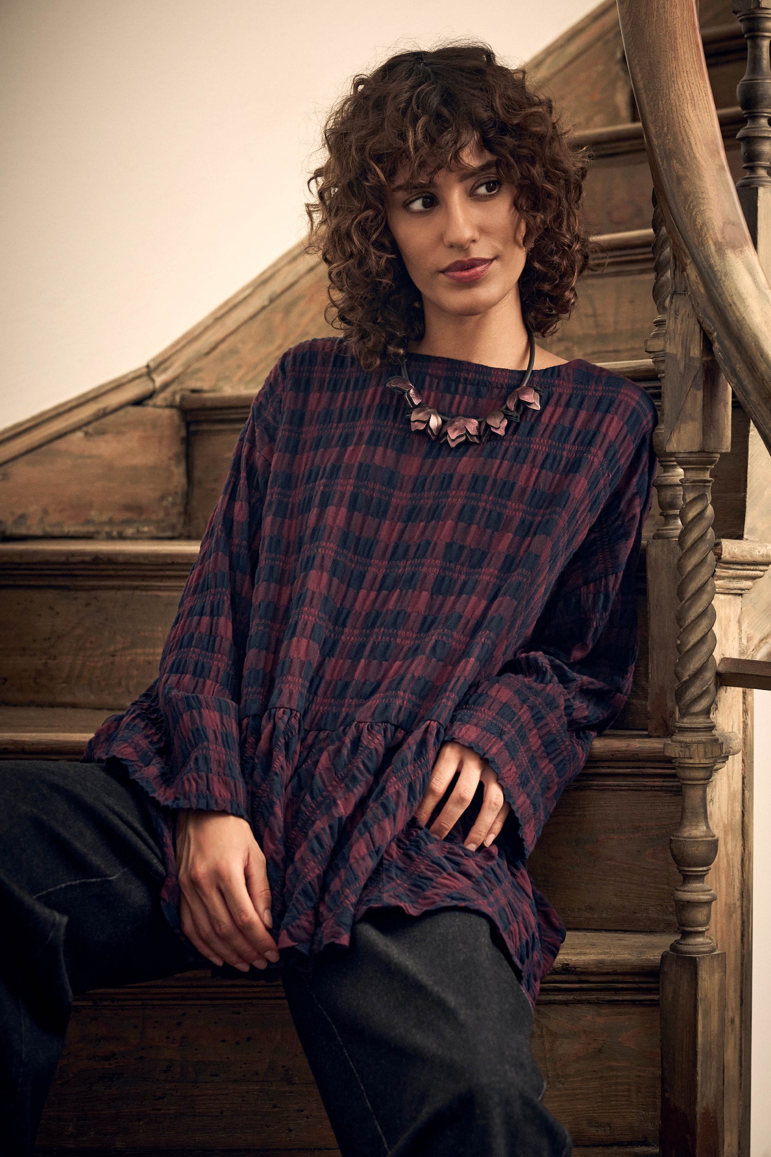 A person with curly hair wearing a maroon and navy blue Crinkle Check Frill Hem Top with a statement necklace, sitting on wooden stairs.