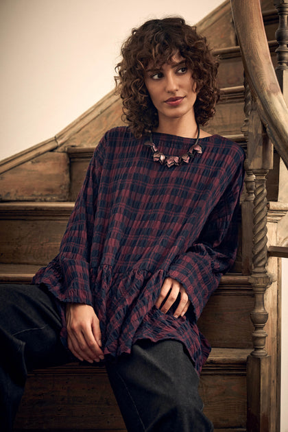 A person with curly hair wearing a maroon and navy blue Crinkle Check Frill Hem Top with a statement necklace, sitting on wooden stairs.