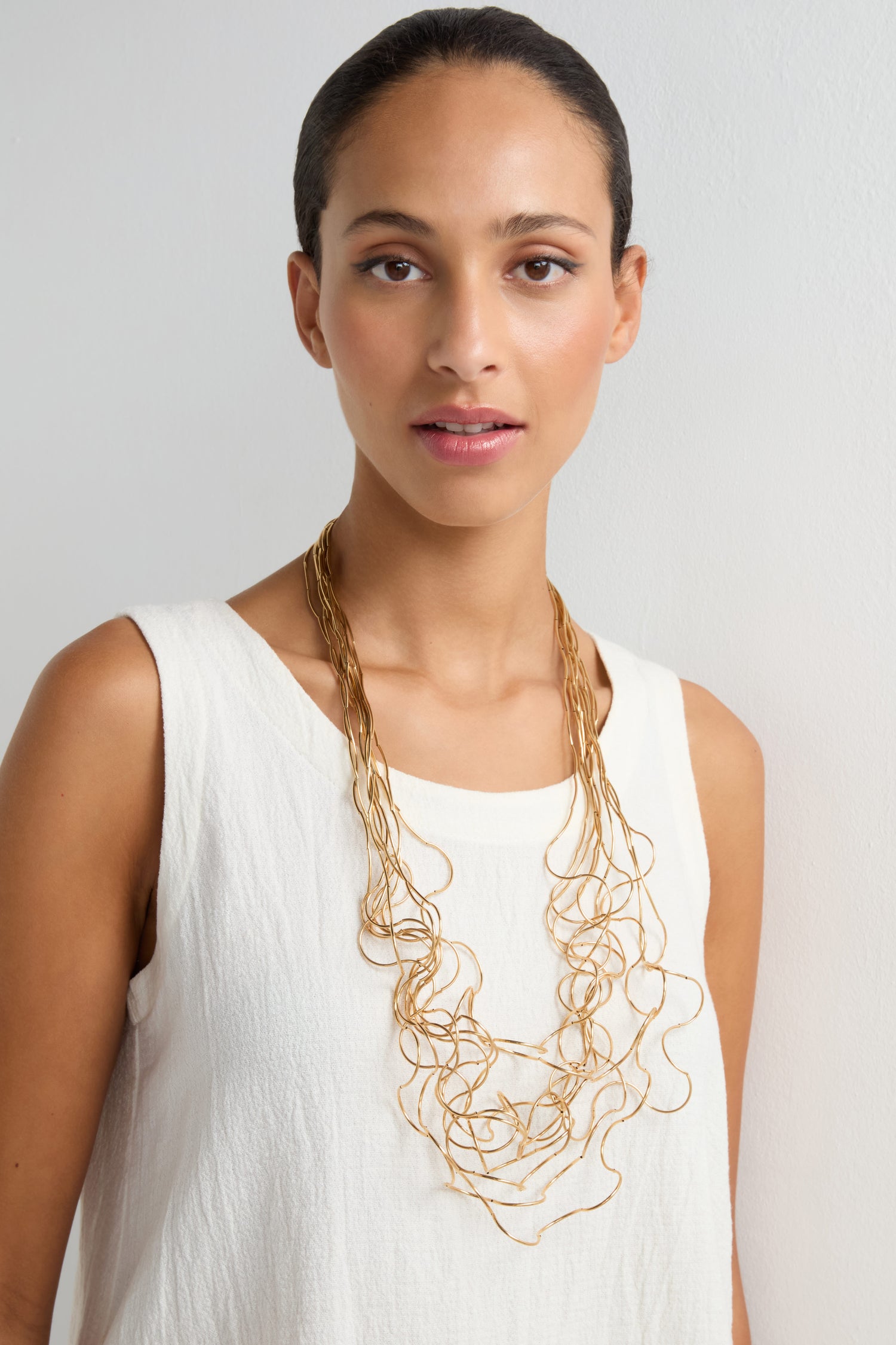 A woman in a sleeveless white top models the Long Medley Necklace—a striking, sculptural accessory—while standing against a plain light backdrop.