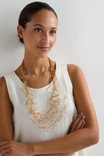 Woman with pulled-back hair wearing a sleeveless white top and the Long Medley Necklace, noted for its sculptural elegance, standing against a plain white wall.