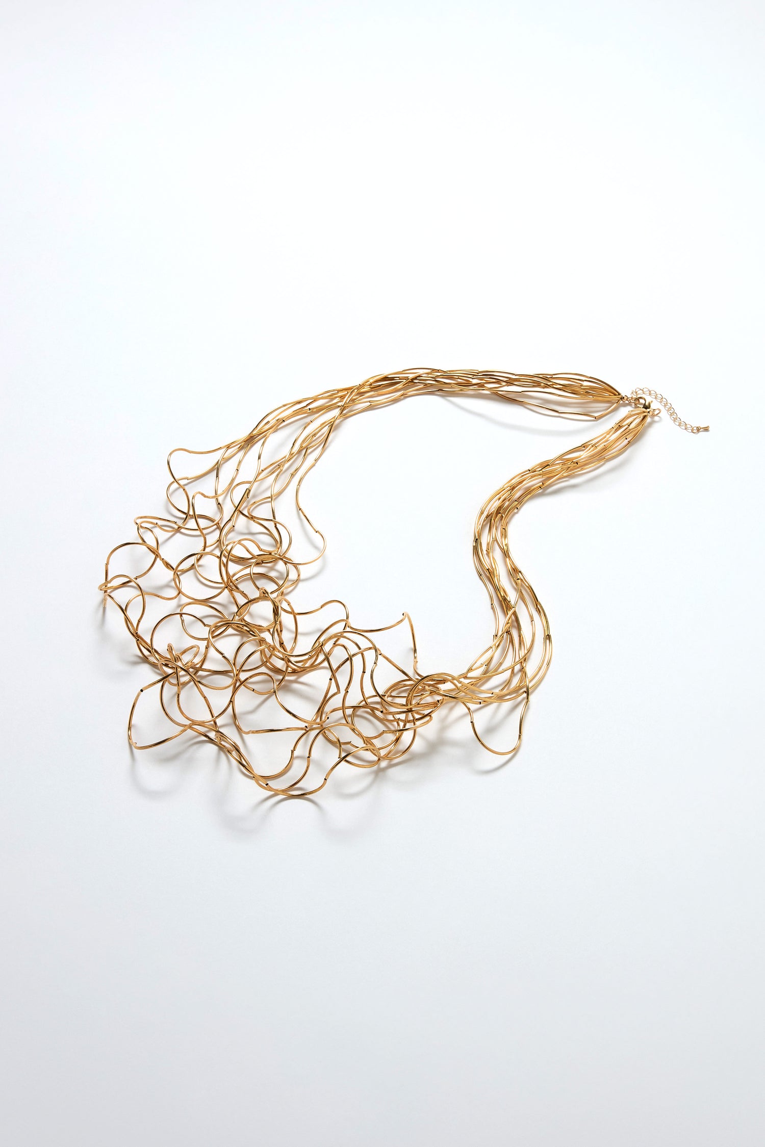 The Long Medley Necklace is a gold-toned metallic piece with sculptural elegance, featuring intertwined multi-strand construction in an abstract, tangled design displayed on a white background.