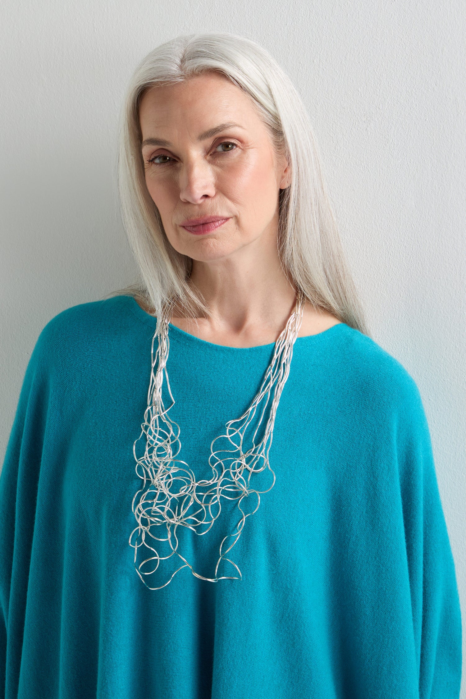 A woman with long gray hair, dressed in a turquoise top and wearing the Long Medley Necklace, stands against a light-colored wall.