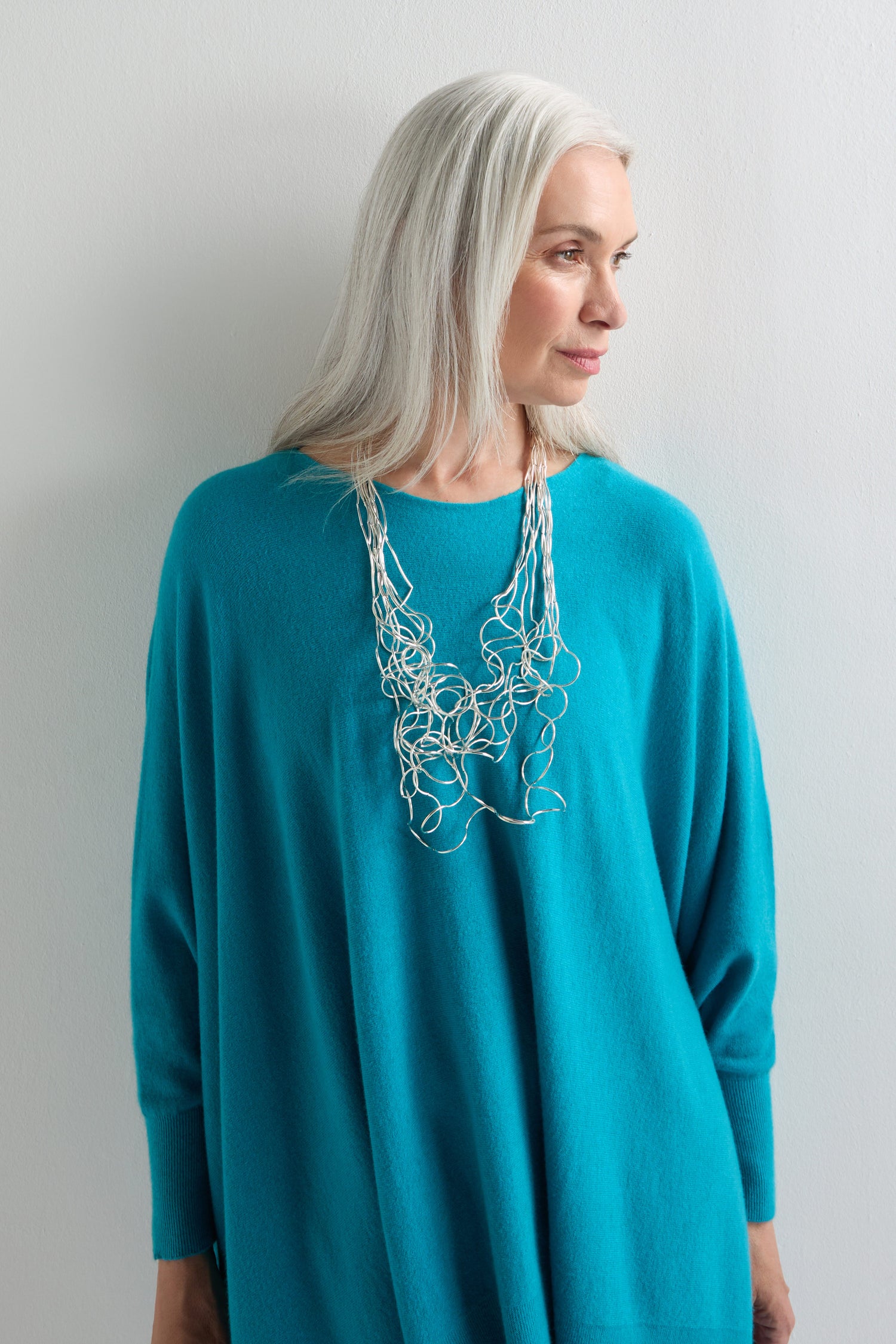 A woman with long gray hair, dressed in a loose turquoise sweater, models the sculptural Long Medley Necklace as she stands against a plain white wall.