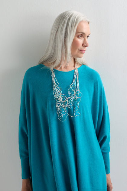 A woman with long gray hair, dressed in a loose turquoise sweater, models the sculptural Long Medley Necklace as she stands against a plain white wall.