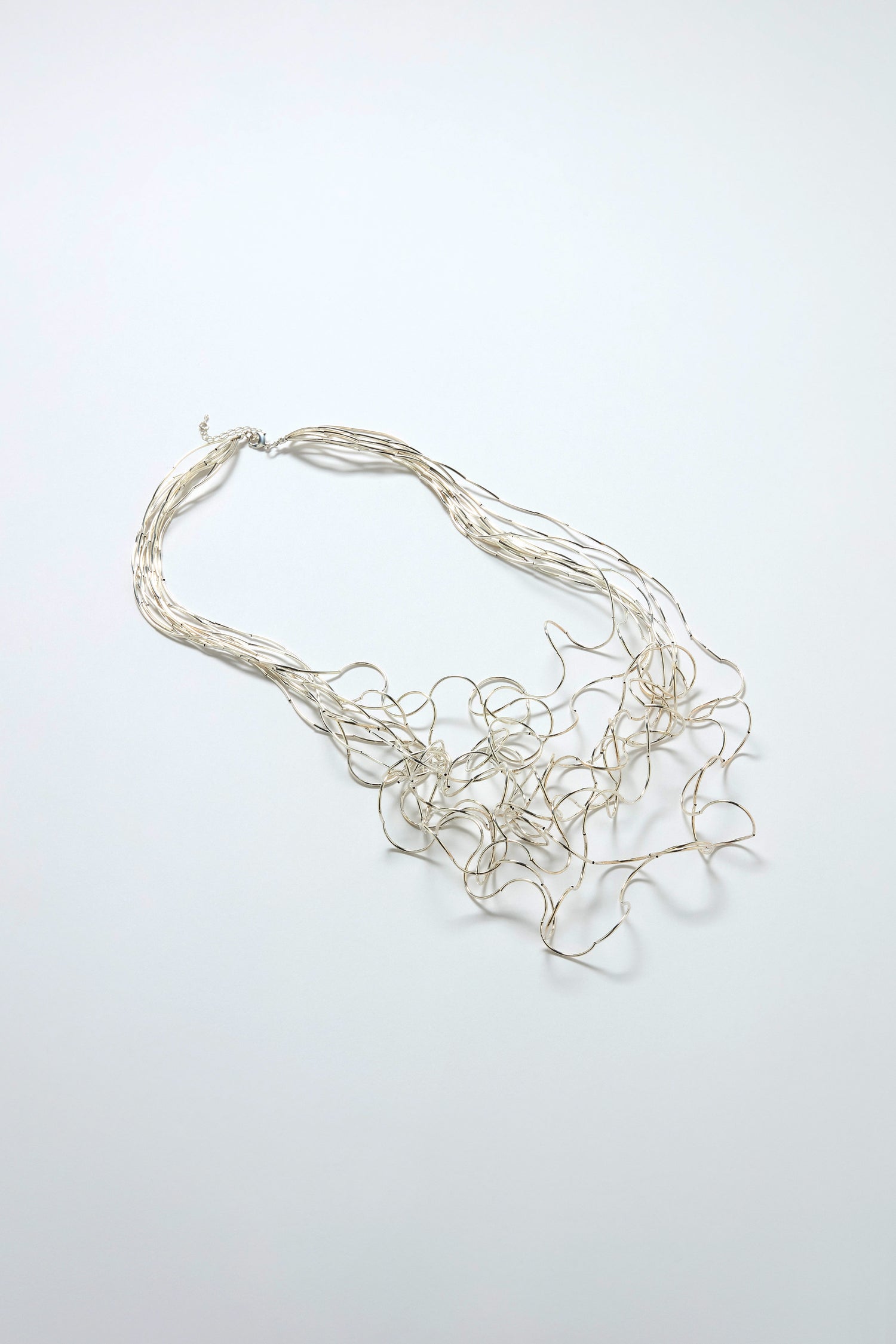 The Long Medley Necklace features a metallic, multi-strand design with thin, wavy strands arranged in an abstract, tangled pattern, shown against a plain light background.