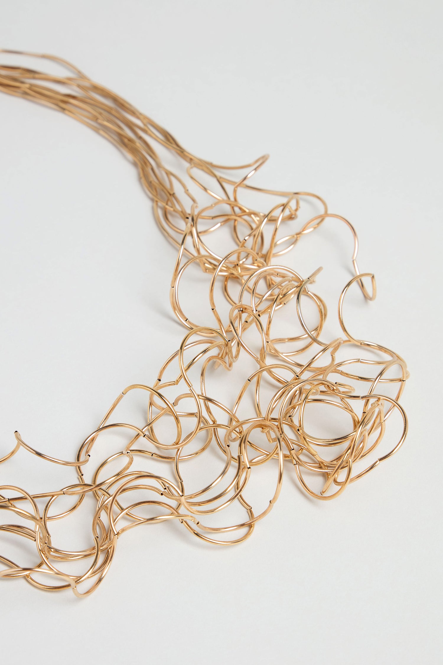 The Gold Long Medley Necklace rests on a white surface, its intricate overlapping loops and abstract design making it an unmistakable statement piece that brings bold elegance to any ensemble.