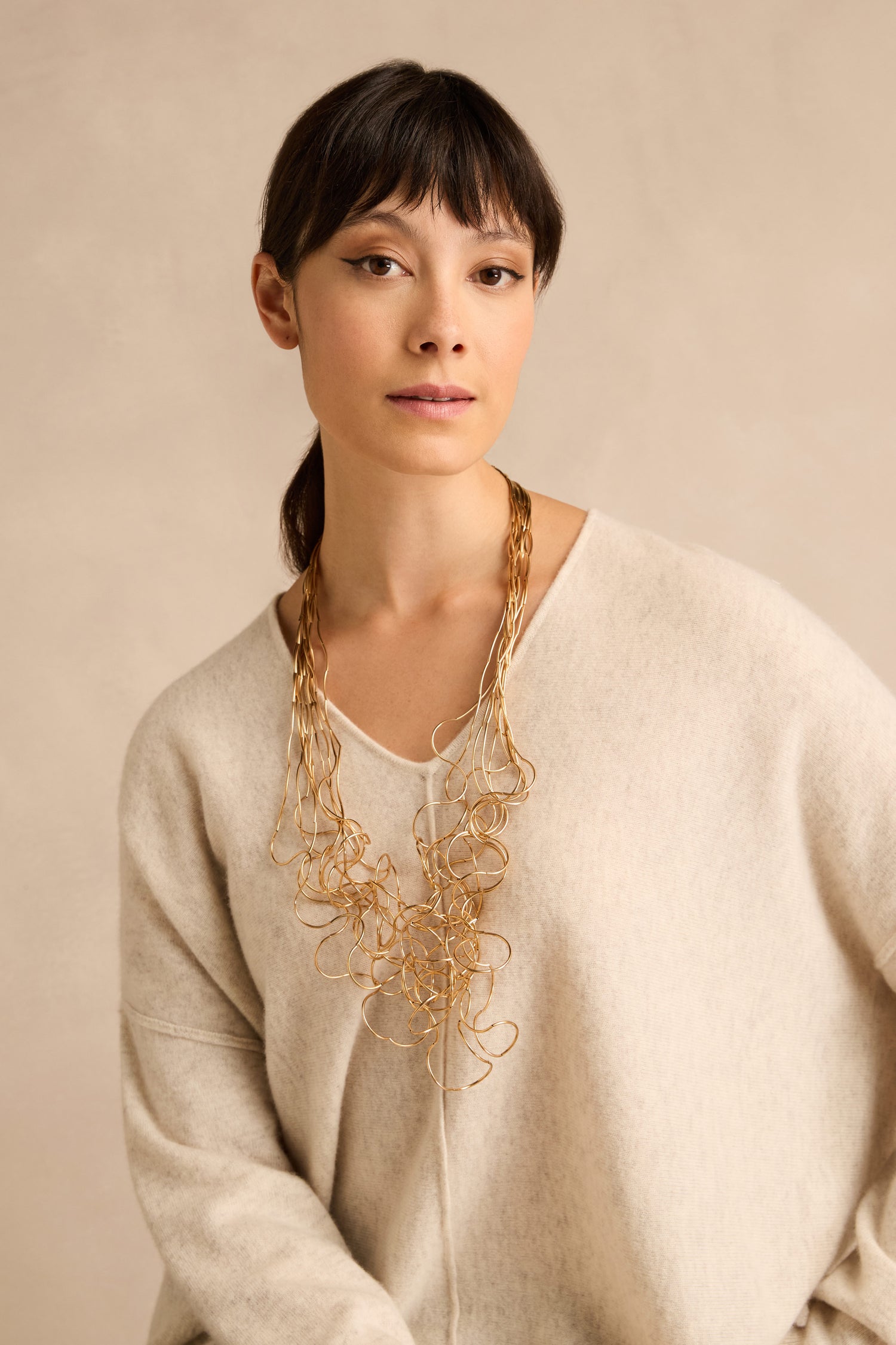Woman with straight dark hair, styled in a cream sweater and the Gold Long Medley Necklace, posed against a neutral beige background.