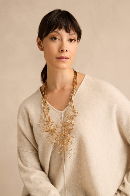 Woman with straight dark hair, styled in a cream sweater and the Gold Long Medley Necklace, posed against a neutral beige background.