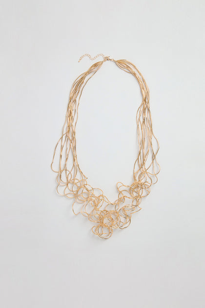 The Gold Long Medley Necklace features several thin, twisted gold chains forming a clustered, abstract pattern at the center, beautifully displayed against a plain light background.