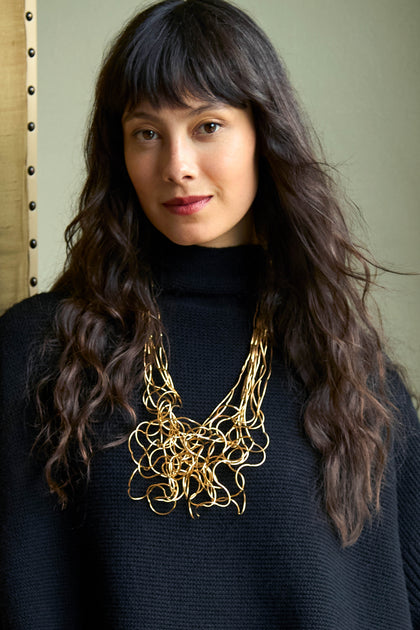 A person wearing a black turtleneck sweater and a Gold Long Medley Necklace featuring a cluster of interconnected wire loops.