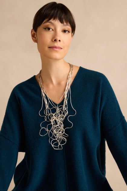 Woman with short dark hair wearing a blue V-neck sweater and the Silver Long Medley Necklace, posed in front of a beige background.