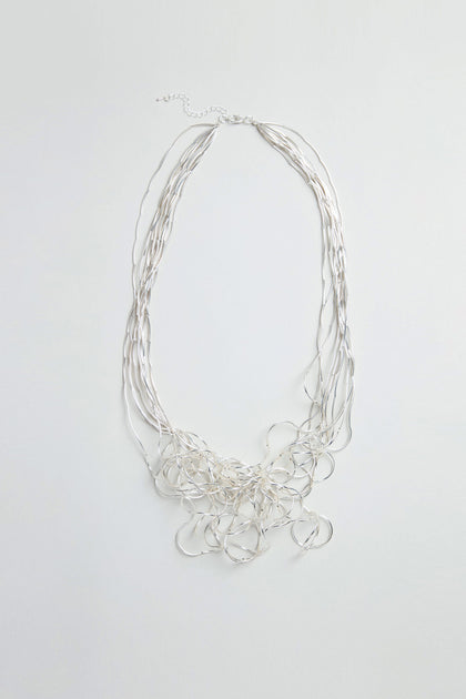 The Silver Long Medley Necklace features multiple strands of thin, irregularly shaped silver loops in an overlapping wire design and is elegantly showcased against a plain white background.