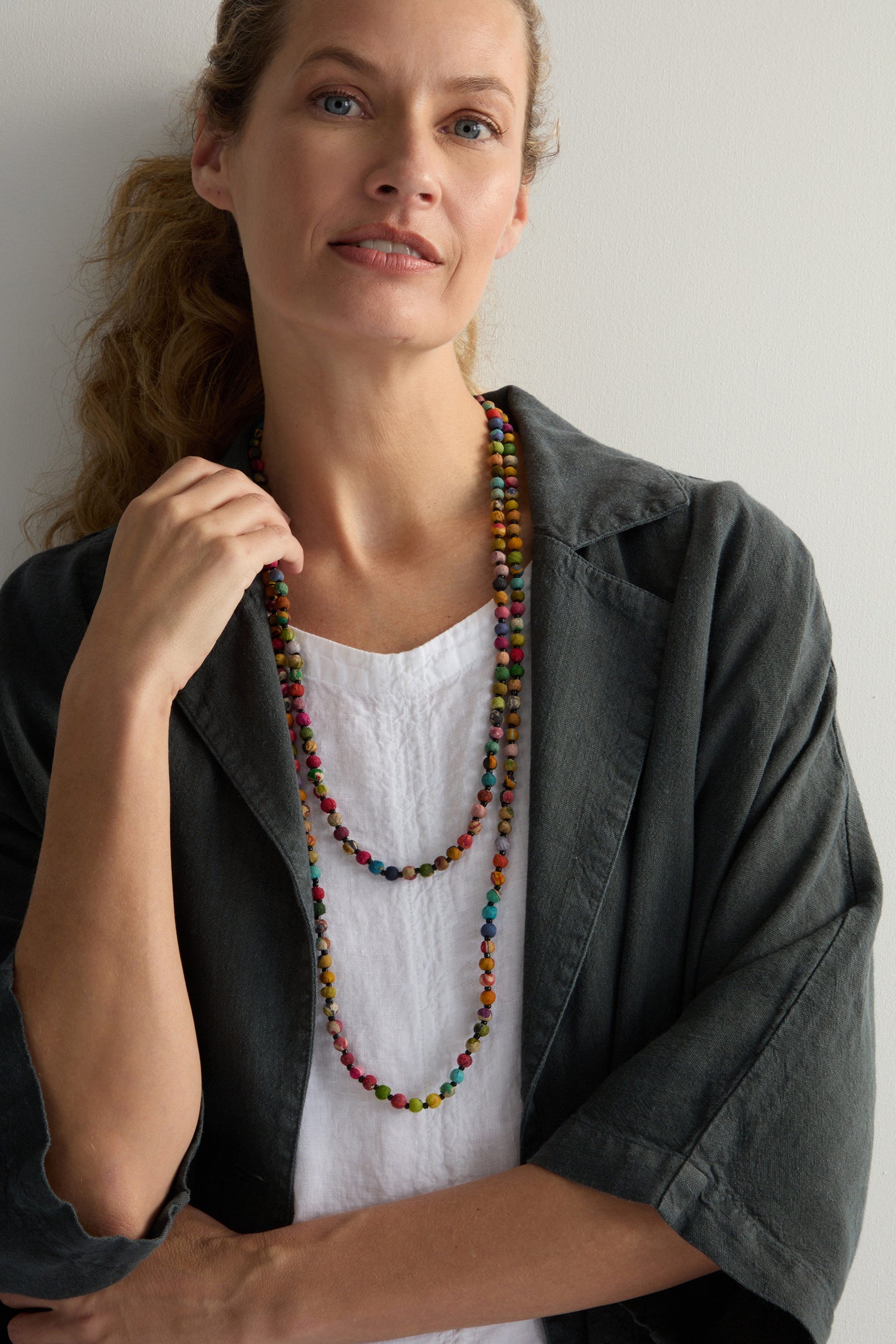 A person stands against a light background in a dark jacket and white top, enhanced by the Kantha Long Bead Spheres Necklace, featuring colorful, hand-rolled beads made from repurposed textiles.