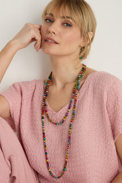 A woman in a textured pink top and pants wears two Kantha Long Bead Spheres Necklaces. She sits with her hand resting against her face, looking at the camera.