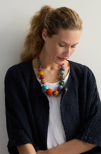 A woman with a ponytail, wearing a textured black jacket, white shirt, and a Kantha Graduated Bead Necklace made from recycled sari fabric, looks down against a plain backdrop.