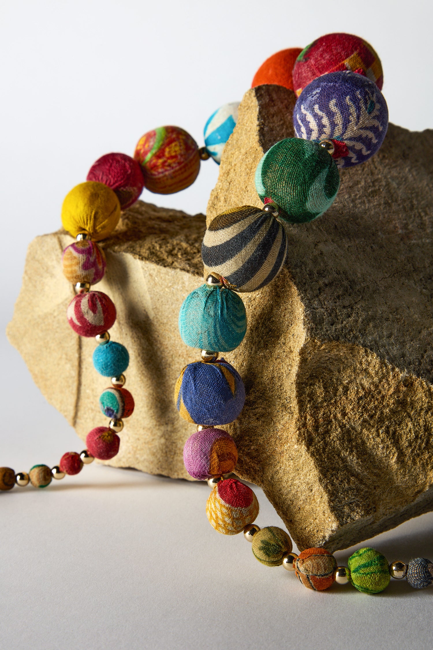 A vibrant Kantha Graduated Bead Necklace, crafted from recycled sari fabric beads, elegantly adorns a textured rock on a white surface.