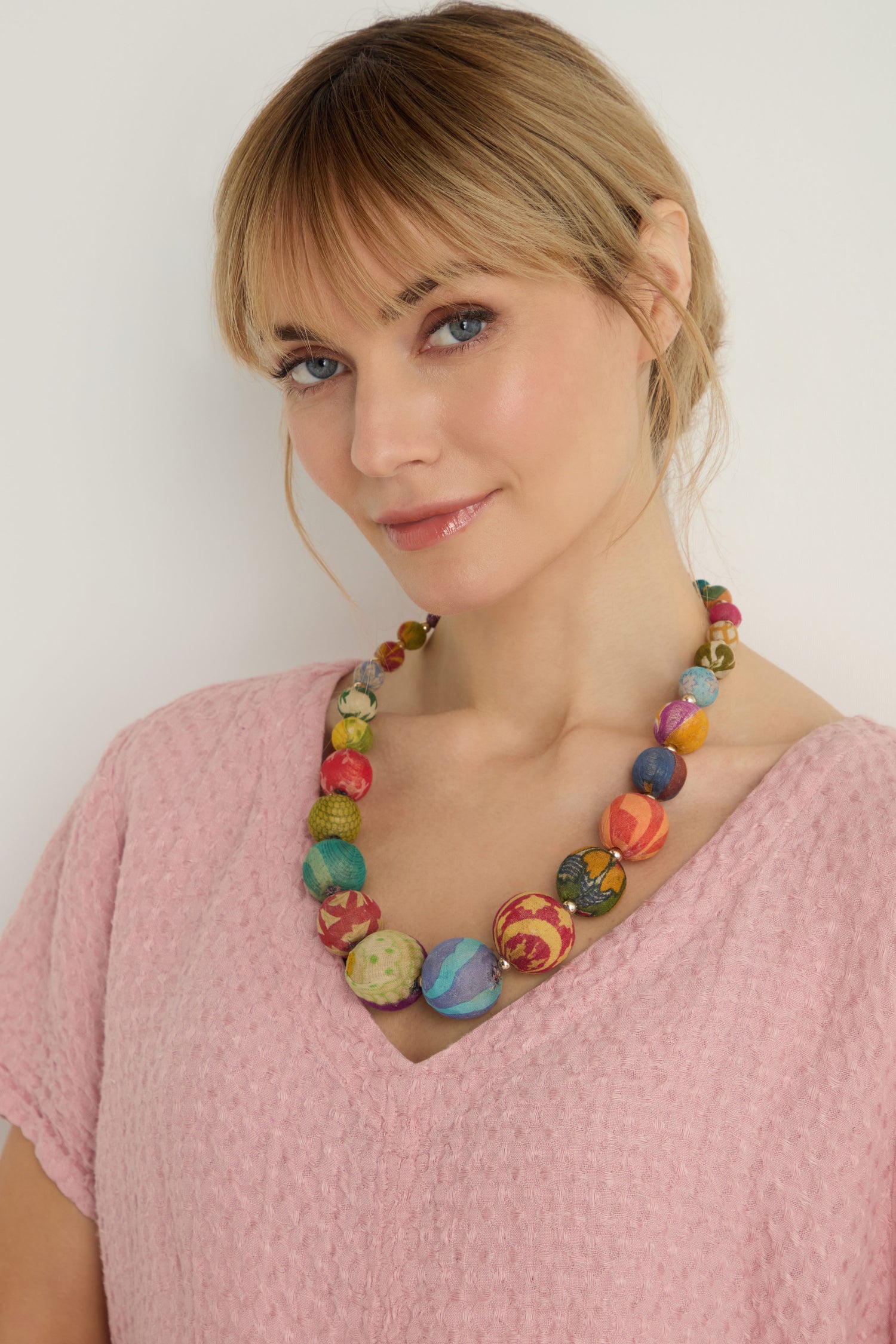 A woman wearing a light pink textured top and the Kantha Graduated Bead Necklace, handcrafted from recycled cotton beads, stands against a plain light background.