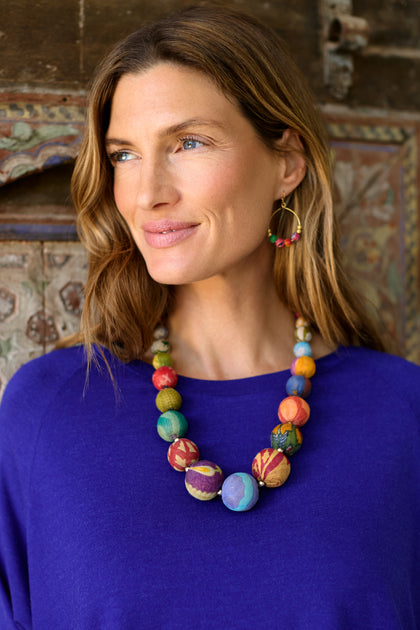 Woman wears a blue top, Kantha Graduated Bead Necklace, and matching hoop earrings against a patterned background, looking to the side—an eye-catching look.
