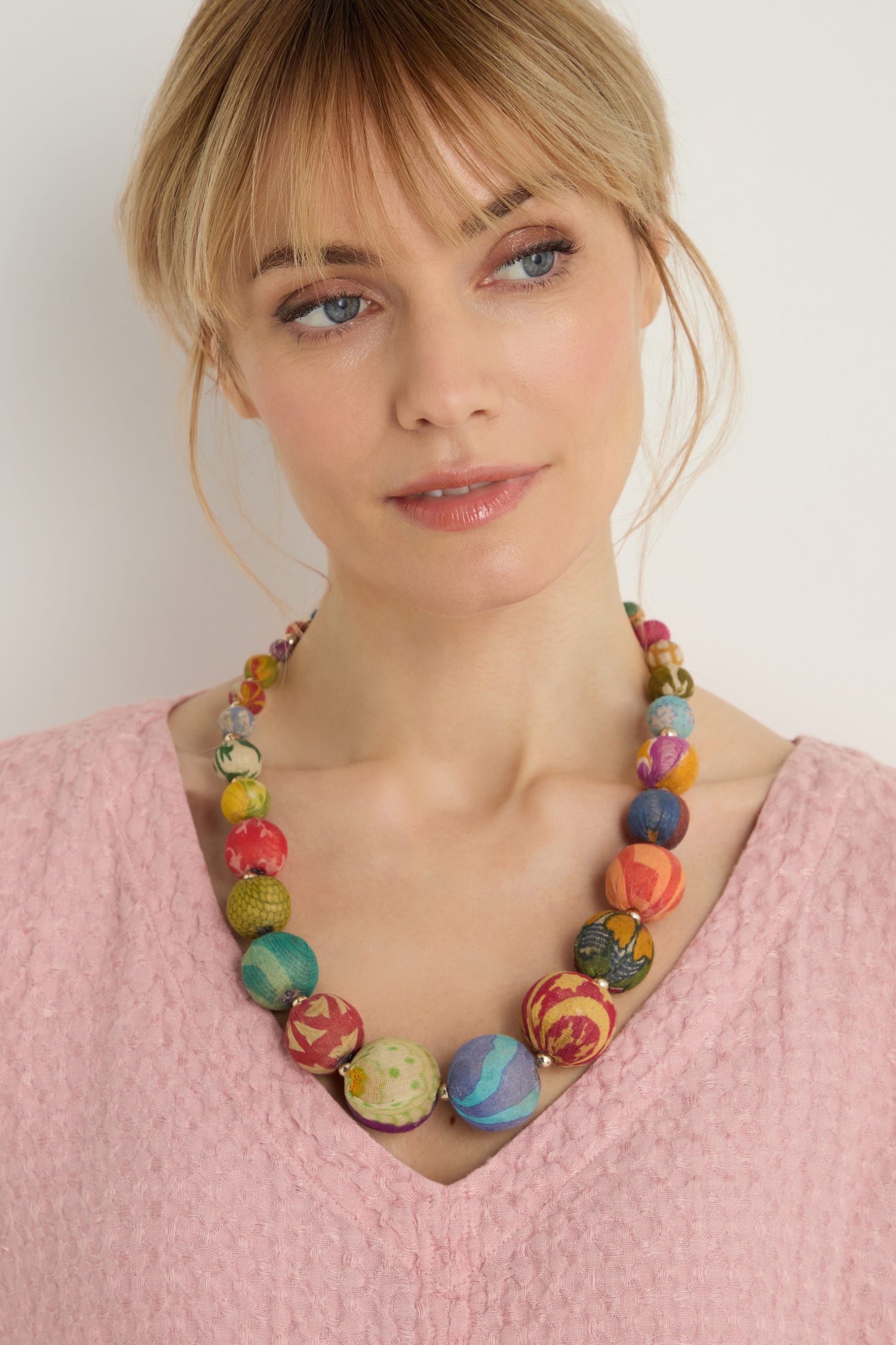A woman in a textured light pink top wears the Kantha Graduated Bead Necklace, handcrafted from recycled cotton beads, as she gazes slightly to the side against a plain background.