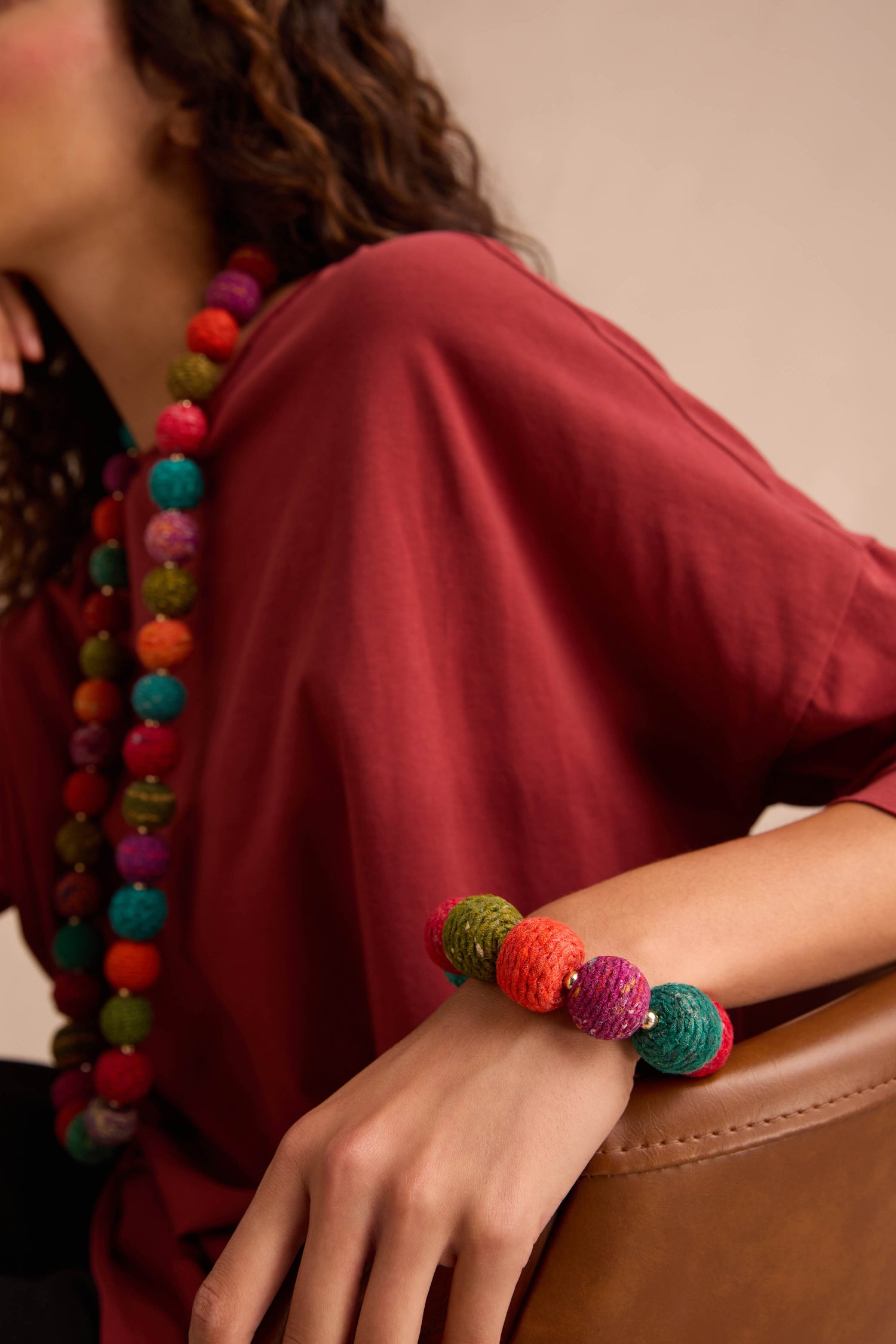 Person wearing a red shirt, colorful cotton-wrapped wooden beads necklace, and the Rainbow Kantha Spheres Bracelet, resting their arm on a brown chair.