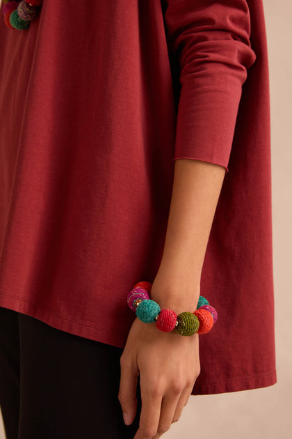 A person wearing a red long-sleeve shirt and black pants, with the Rainbow Kantha Spheres Bracelet—featuring cotton-wrapped wooden beads—adorning their wrist.