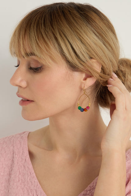 Woman with blonde hair in a low bun, wearing a pink textured top and Kantha Gilded Hoop Earrings with colorful beads, touches her hair while facing left.