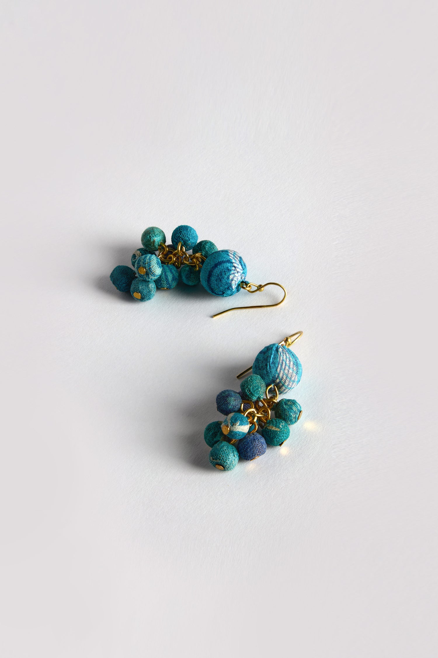 Two Kantha Spheres Earrings with teardrop shapes, adorned in turquoise and blue fabric beads crafted from recycled textiles on gold hooks, rest on a white surface.