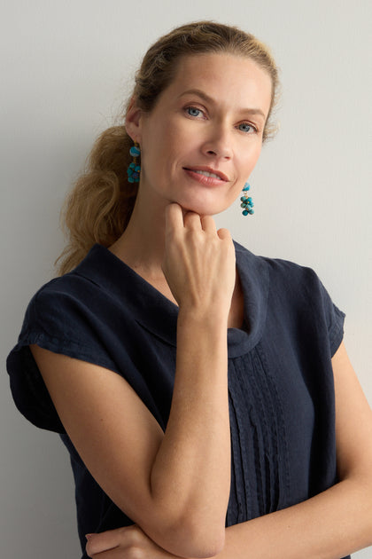 A woman with long hair in a black dress thoughtfully poses with her hand under her chin, showcasing Kantha Spheres Earrings. These earrings are crafted from recycled textiles, adding a vibrant blue touch against the plain background.