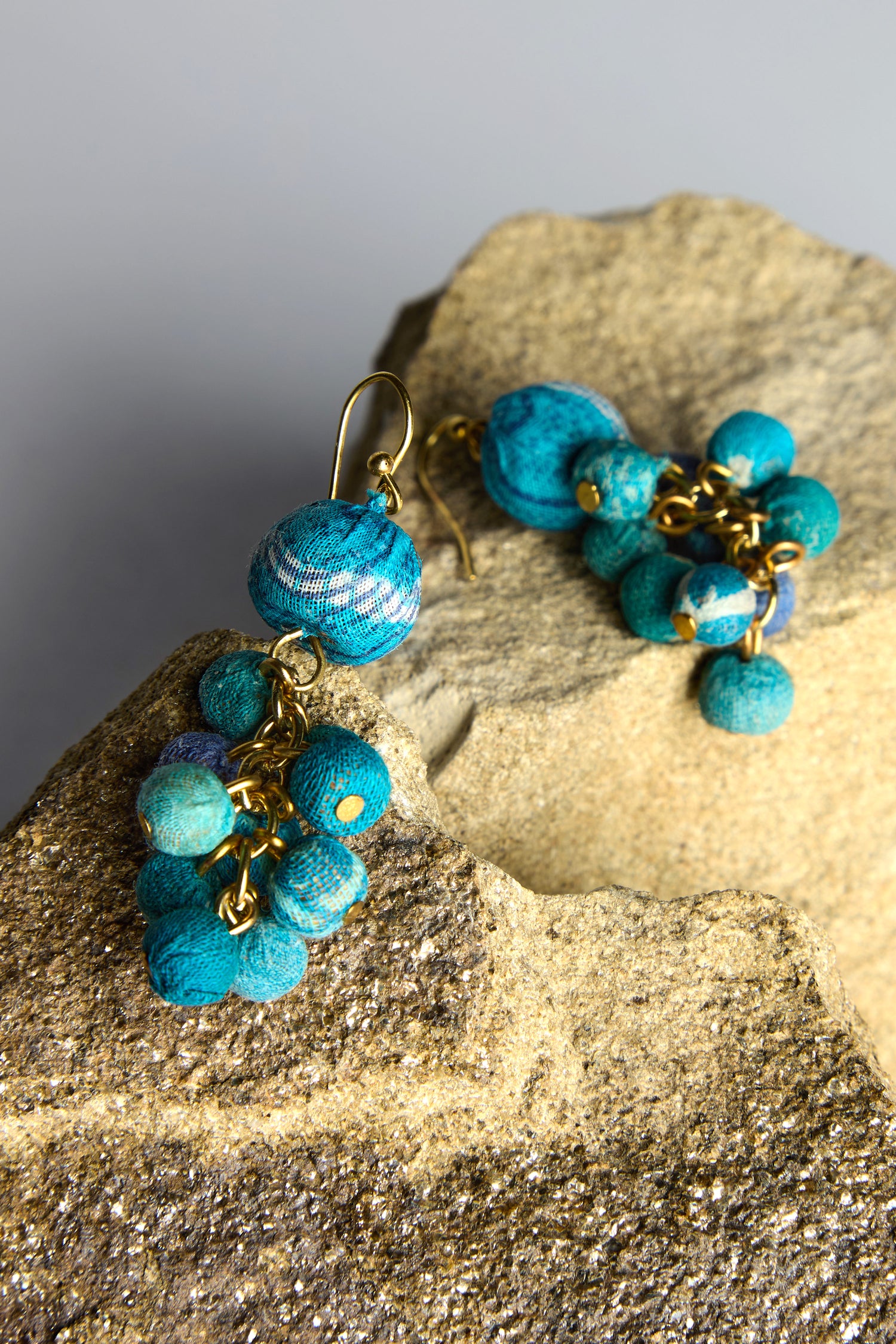 Introducing the Kantha Spheres Earrings, featuring dangling blue and gold beads made from recycled textiles. Lovingly handcrafted, they exhibit rustic charm and vibrant allure on rough stone surfaces.