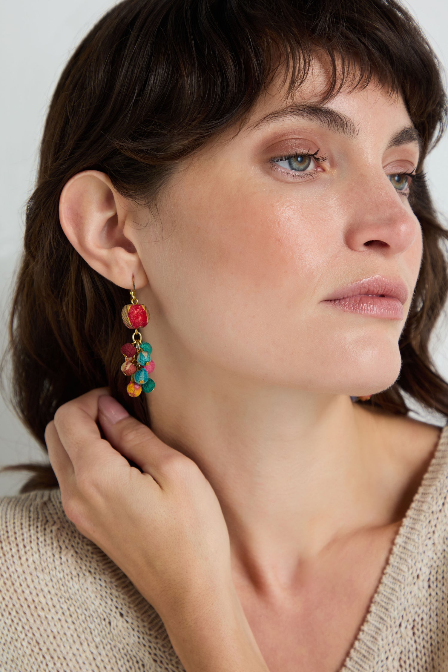 A woman with light skin and short brown hair wears a beige knit top and Kantha Spheres Earrings—colorful handcrafted fabric bead earrings—while gazing to the side and touching her neck.