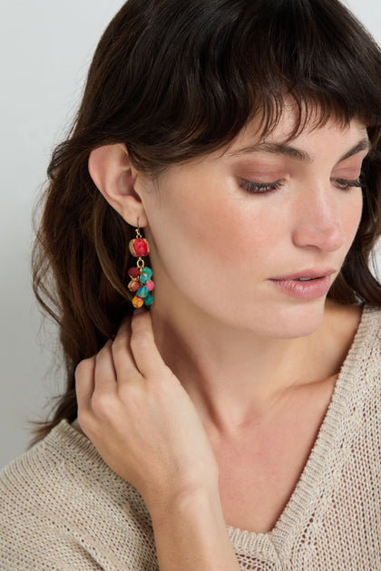 A woman with brown hair wears a knit beige top and touches her Kantha Spheres Earrings—colorful, handcrafted earrings made from fabric beads—while looking down.