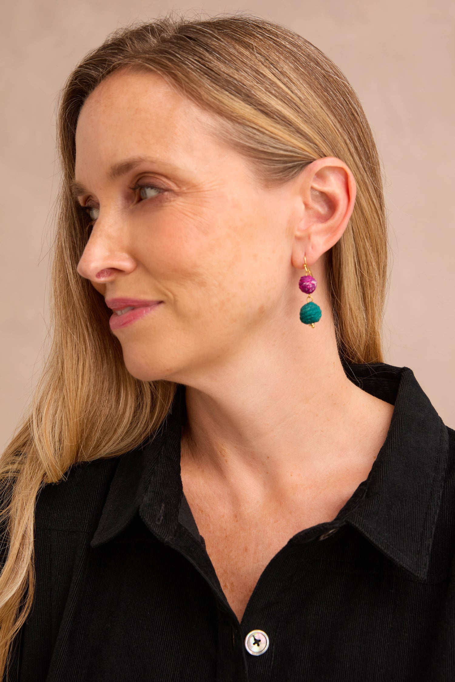 A woman with long blonde hair, dressed in a black collared shirt, showcases the Kantha Double Spheres Earrings—vibrant statement pieces featuring cotton-wrapped Kantha beads—as she looks to the left against a plain background.