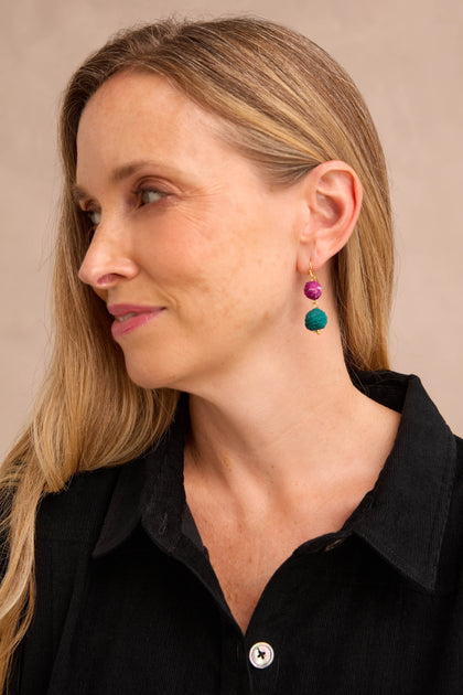 A woman with long blonde hair, dressed in a black collared shirt, showcases the Kantha Double Spheres Earrings—vibrant statement pieces featuring cotton-wrapped Kantha beads—as she looks to the left against a plain background.