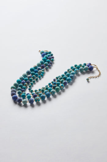 The Kantha Cascade Necklace is a multi-strand piece with round, blue-green beads made from repurposed fabric and gold accents, set on a light gray background. It highlights sustainable fashion and includes an adjustable chain closure for a custom fit.